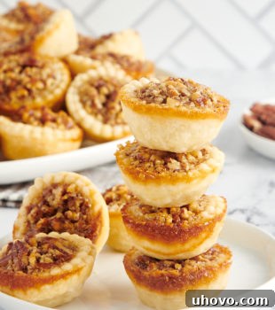 A stack of four mini pecan pies on a plate next to two other mini pies, showcasing their golden-brown crust and glistening pecan topping.
