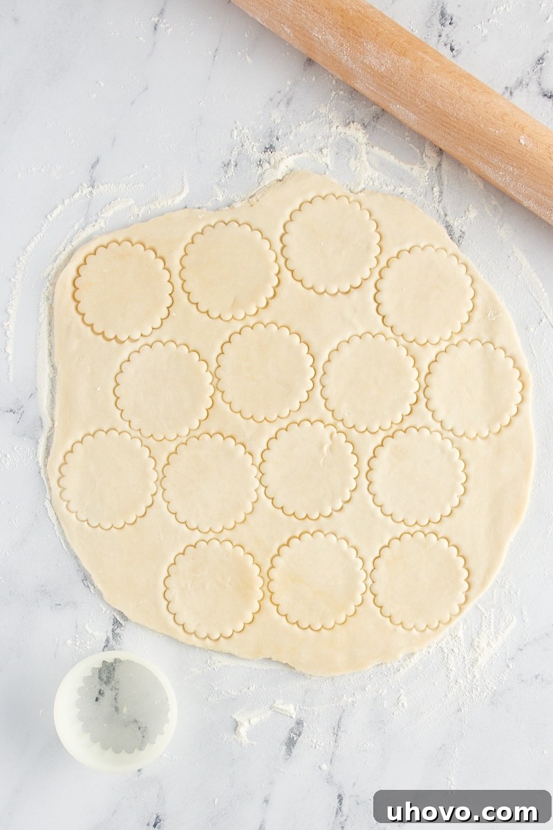 A hand cutting small, perfectly round circles of pie crust from a larger rolled-out pastry using a cookie cutter.