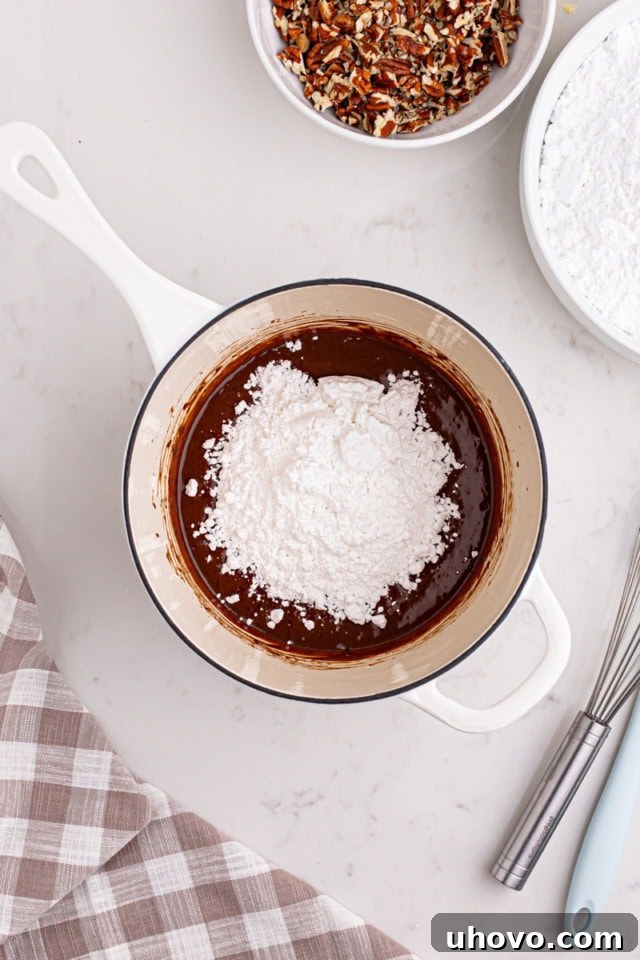 Confectioners' sugar and vanilla extract being added to the chocolate and milk mixture in a saucepan, ready to be whisked.