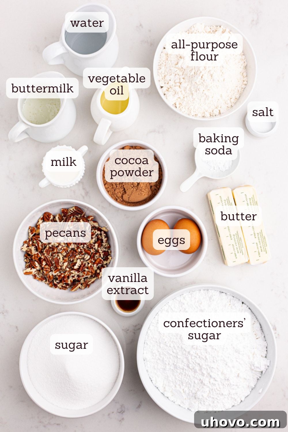 Overhead view of various key ingredients for Texas Sheet Cake laid out on a marble surface, including flour, sugar, butter, buttermilk, cocoa powder, and eggs.