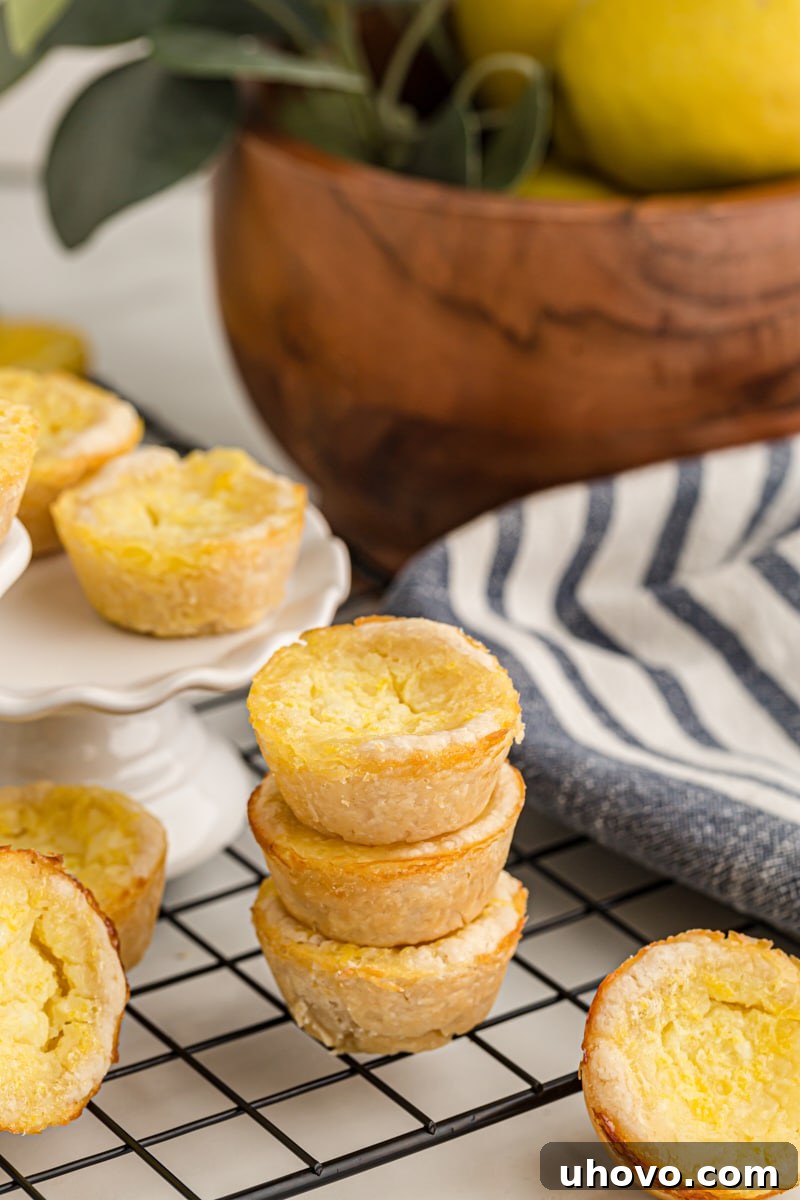 A small stack of three golden Lemon Chess Tartlets on a wire rack, with more tartlets blurred in the background, ready to be devoured.