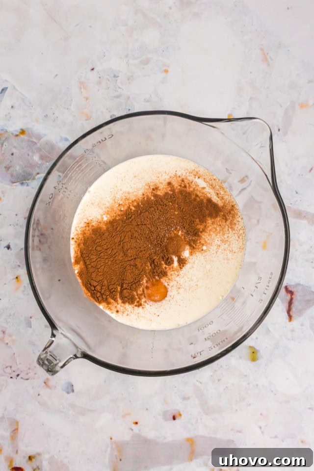 An overhead shot of various custard ingredients – eggs, sugar, vanilla, milk, heavy cream, cinnamon, and salt – meticulously laid out in a mixing bowl before whisking.