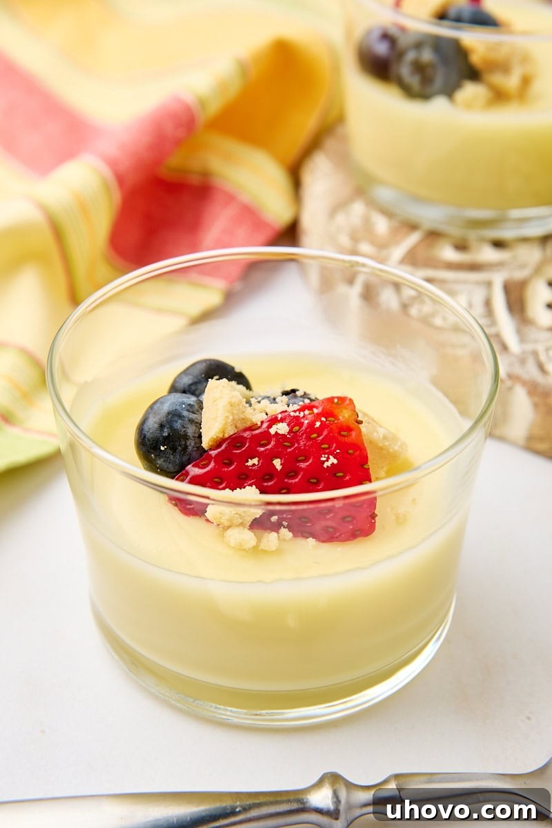 Exquisitely presented lemon posset in clear glass jars, garnished with fresh mixed berries and shortbread crumbles. More servings are visible in the soft-focused background, emphasizing its elegant appeal for a gathering.