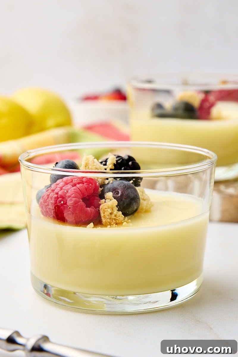 A close-up view of a perfectly set lemon posset, topped generously with glistening fresh berries and crumbled shortbread, served in a transparent glass, with more delicious servings blurred in the background.