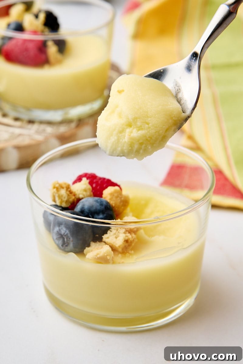 A close-up shot capturing a spoonful of lemon posset, creamy and perfectly set, hovering enticingly over the remaining portion in a clear glass, highlighting its smooth texture and readiness to be enjoyed.