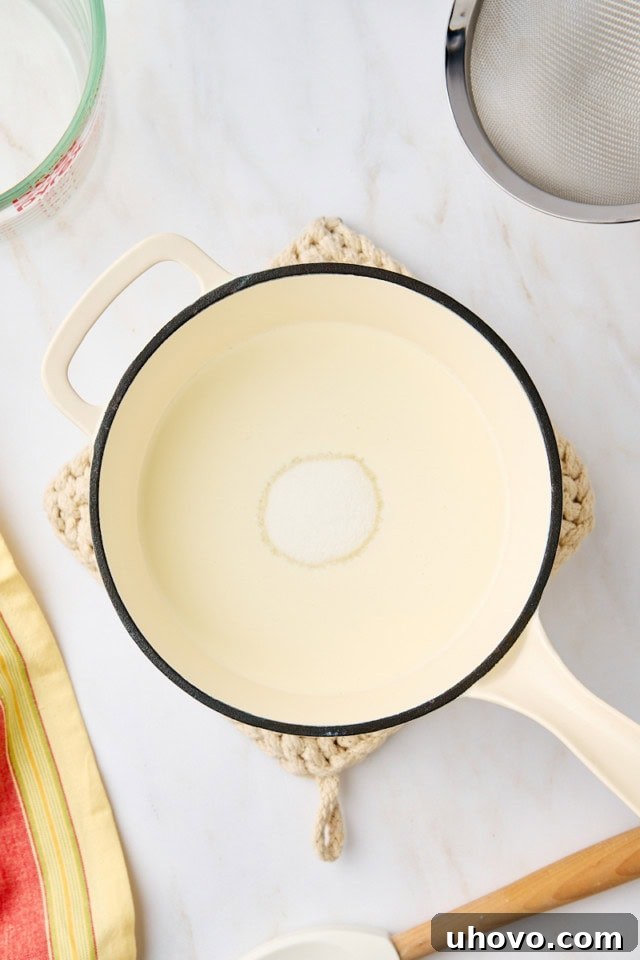 Overhead perspective of heavy cream and granulated sugar combined within a shiny metal saucepan, illuminated by natural light, ready to be heated and stirred on the stovetop.