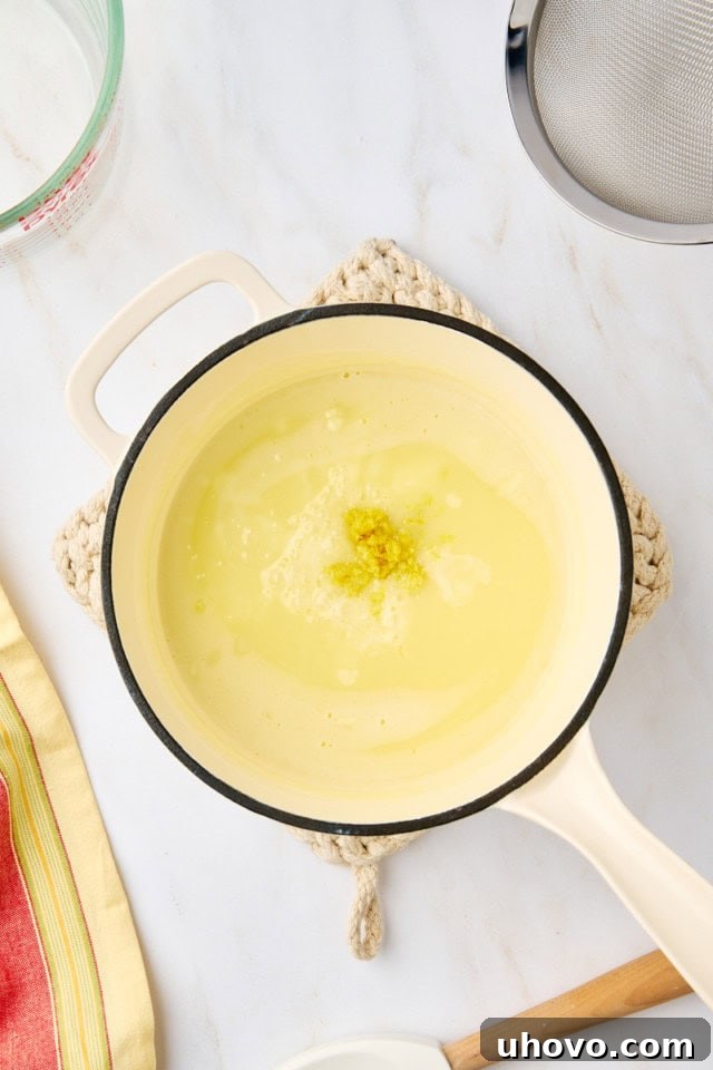 Overhead shot showing freshly squeezed lemon juice and aromatic lemon zest being stirred into the cooked cream and sugar mixture in a saucepan, highlighting the next critical step in making lemon posset.