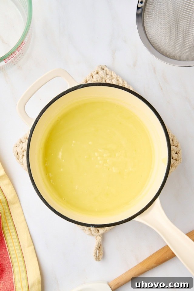 An overhead shot of the lemon posset mixture resting in a saucepan after the lemon juice has been added, showing the liquid starting to cool and slightly thicken, an essential pause before straining.