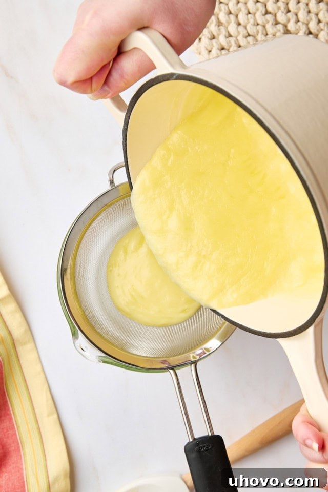 Overhead view of the lemon posset mixture being carefully poured through a fine-mesh sieve into a large, clear glass measuring cup, ensuring a perfectly smooth texture by catching any zest or small curds.