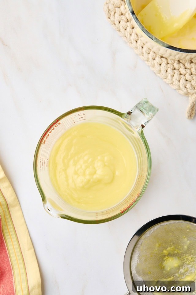 Overhead shot showcasing the perfectly smooth and translucent lemon posset mixture collected in a large, clear glass measuring cup after being strained, ready for individual serving portions.