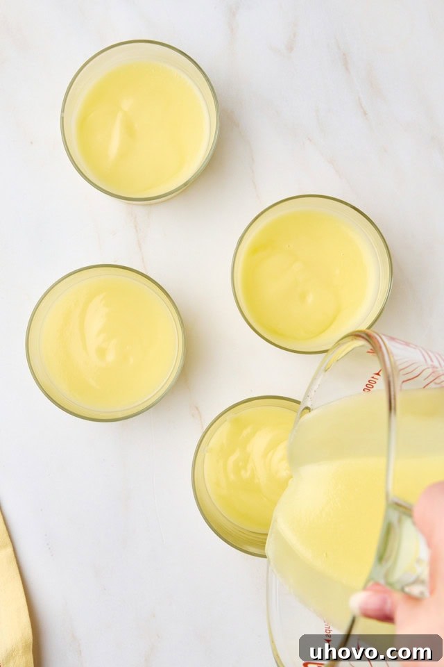 Overhead view of the smooth lemon posset mixture being meticulously poured from a measuring cup into elegant individual serving glasses, indicating the division into portions before chilling.