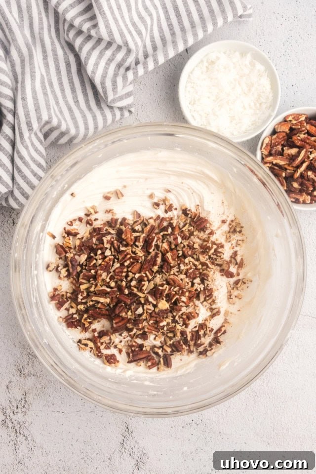 Overhead view of chopped toasted pecans being stirred into a bowl of creamy white cream cheese frosting.