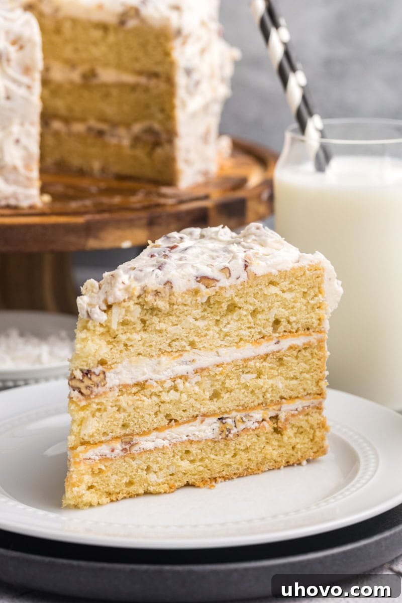 A beautiful slice of Italian cream cake rests on a white plate, with the remainder of the elegantly frosted cake visible in the background.