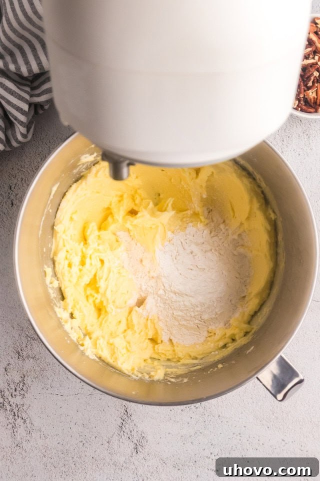Overhead view of dry ingredients being added to the cake batter in the bowl of a stand mixer, with a spatula ready to fold in coconut.