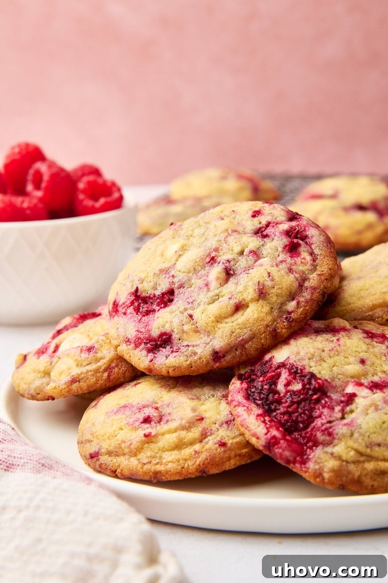 raspberry white chocolate cookies piled on a white plate, with a bowl of raspberries in the background