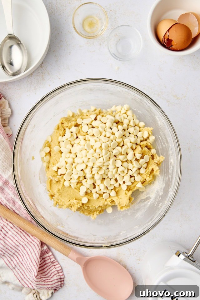 overhead view of white chocolate chips added to cookie dough in a glass mixing bowl