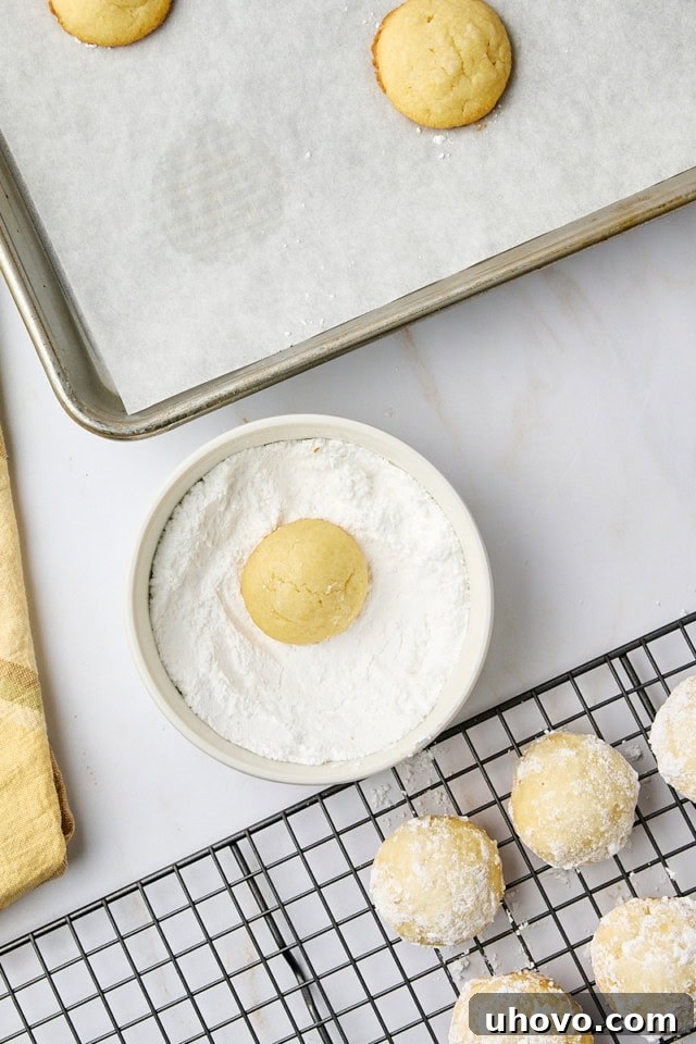 A warm lemon cooler cookie being rolled in a bowl of confectioners' sugar, with more partially coated cookies visible on a wire rack in the background.