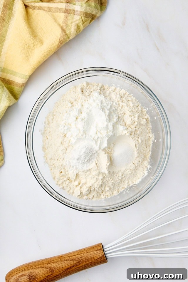 An overhead view of flour, cornstarch, baking powder, and salt precisely measured and combined in a clear glass mixing bowl, ready for the wet ingredients.