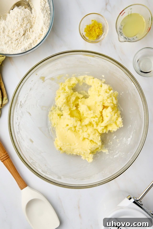 Overhead shot showing fresh lemon zest, tart lemon juice, and optional lemon extract being added to the creamy mixture of butter and sugar in a glass bowl, infusing it with vibrant citrus flavor.