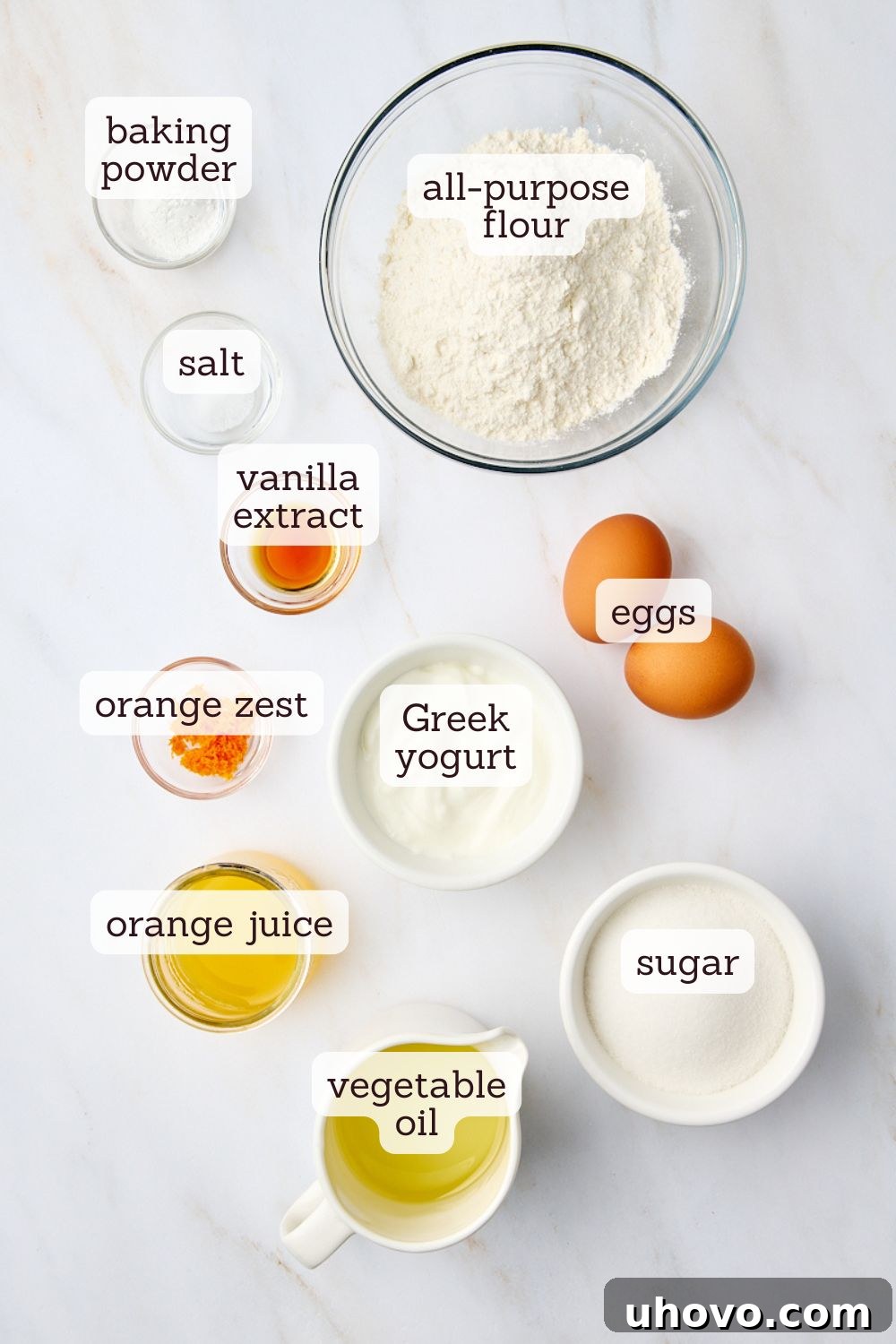 An overhead shot of all the essential ingredients laid out for a vibrant citrus yogurt cake, including flour, sugar, eggs, oil, Greek yogurt, and fresh oranges, ready for baking.
