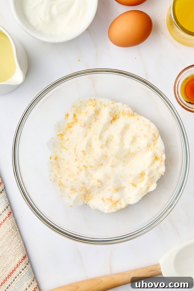 A close-up overhead view of vibrant orange zest mixed with granulated sugar in a glass mixing bowl, illustrating the essential step of rubbing them together to release citrus oils.