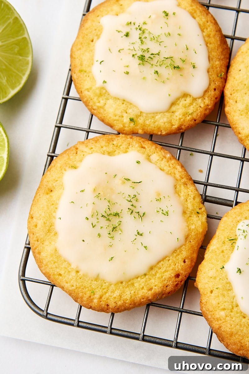Close-up view of a coconut lime cookie on a wire rack surrounded by more cookies and slices of lime, showcasing its soft texture and zesty glaze.