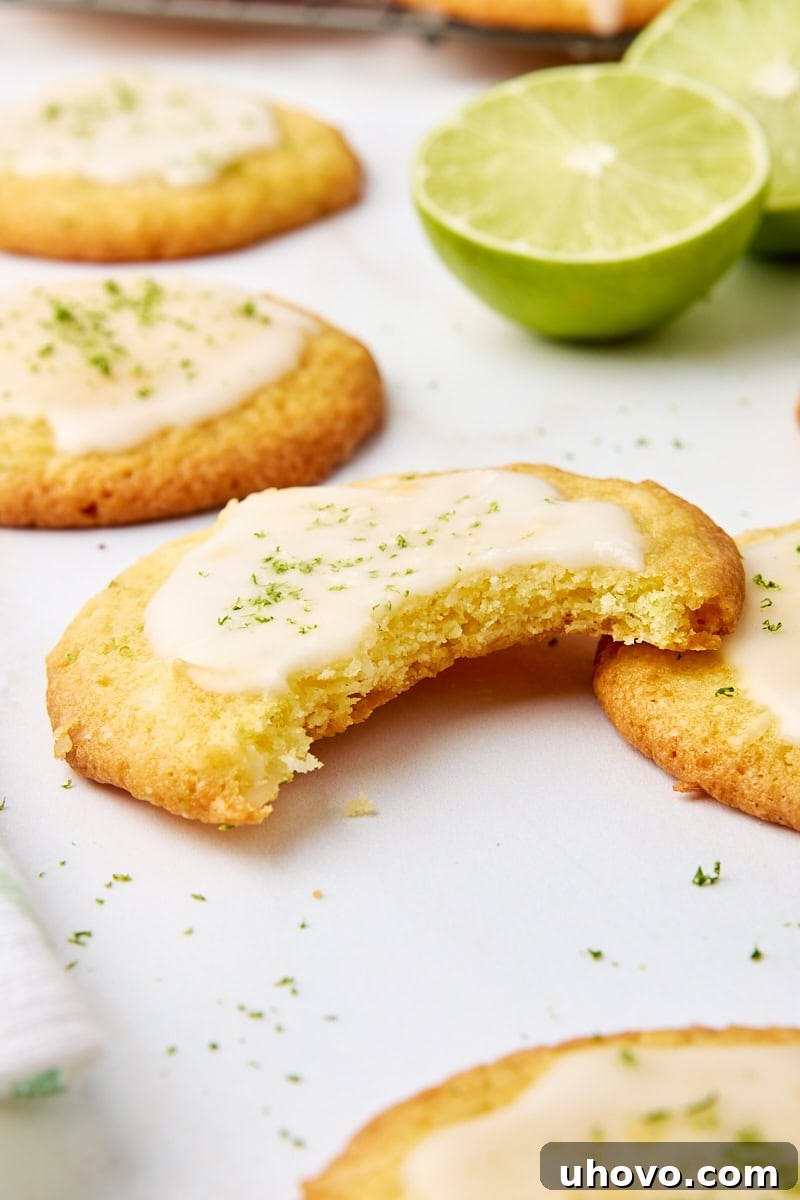 A coconut lime cookie with a bite taken out, showcasing its soft interior, surrounded by whole cookies and fresh lime slices on a white surface.