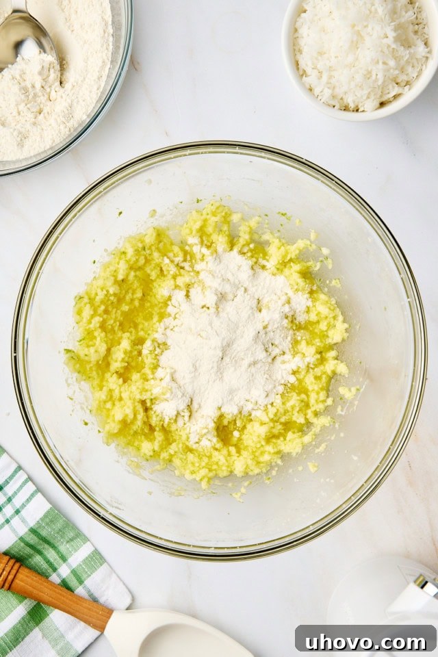 Overhead view of sweetened shredded coconut being gently folded into the prepared cookie dough in a glass mixing bowl.