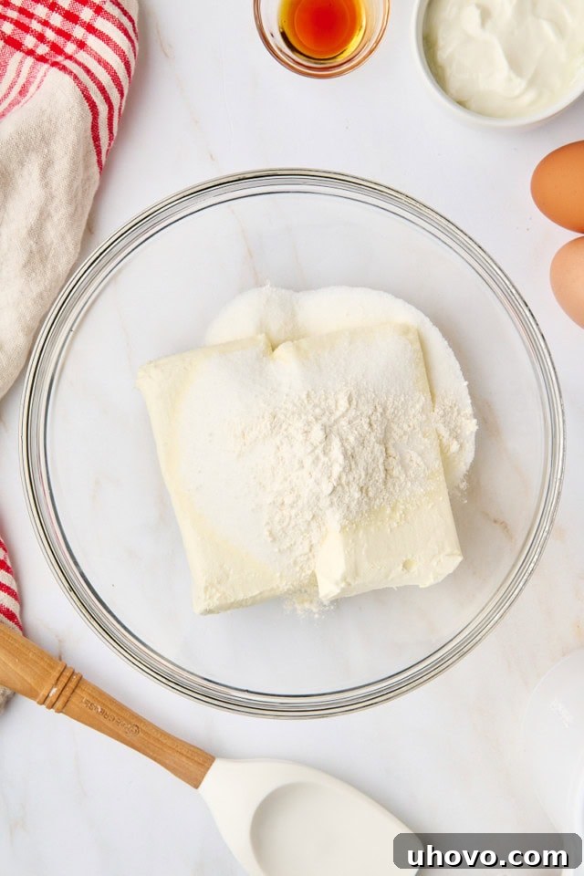 Cream cheese, granulated sugar, and all-purpose flour being beaten together in a glass mixing bowl with an electric mixer for the cheesecake filling.