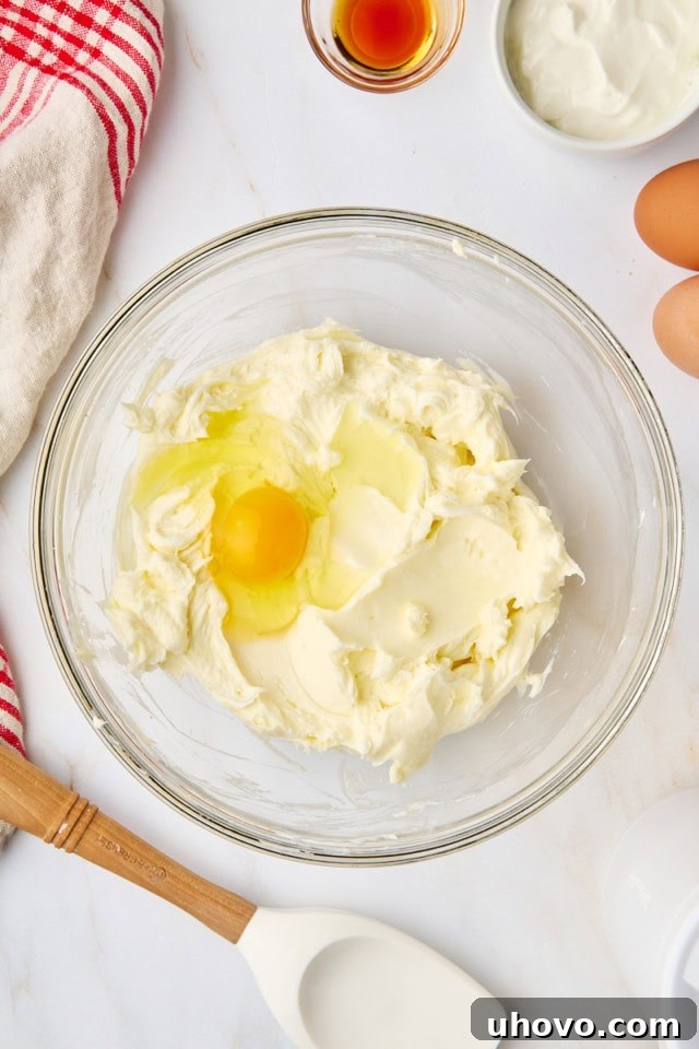 An egg being carefully cracked and added to the creamy, beaten cream cheese and sugar mixture in a glass bowl.
