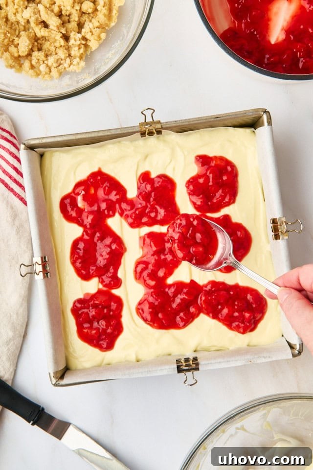 Luscious strawberry topping being carefully spooned and spread evenly over the creamy cheesecake layer in the baking pan.