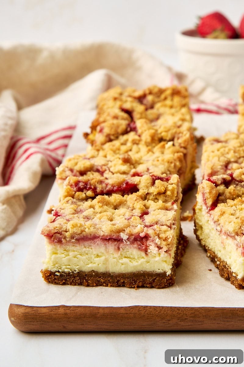 A single strawberry cheesecake bar on parchment paper, surrounded by more delicious bars and a bowl of fresh, ripe strawberries in the background.