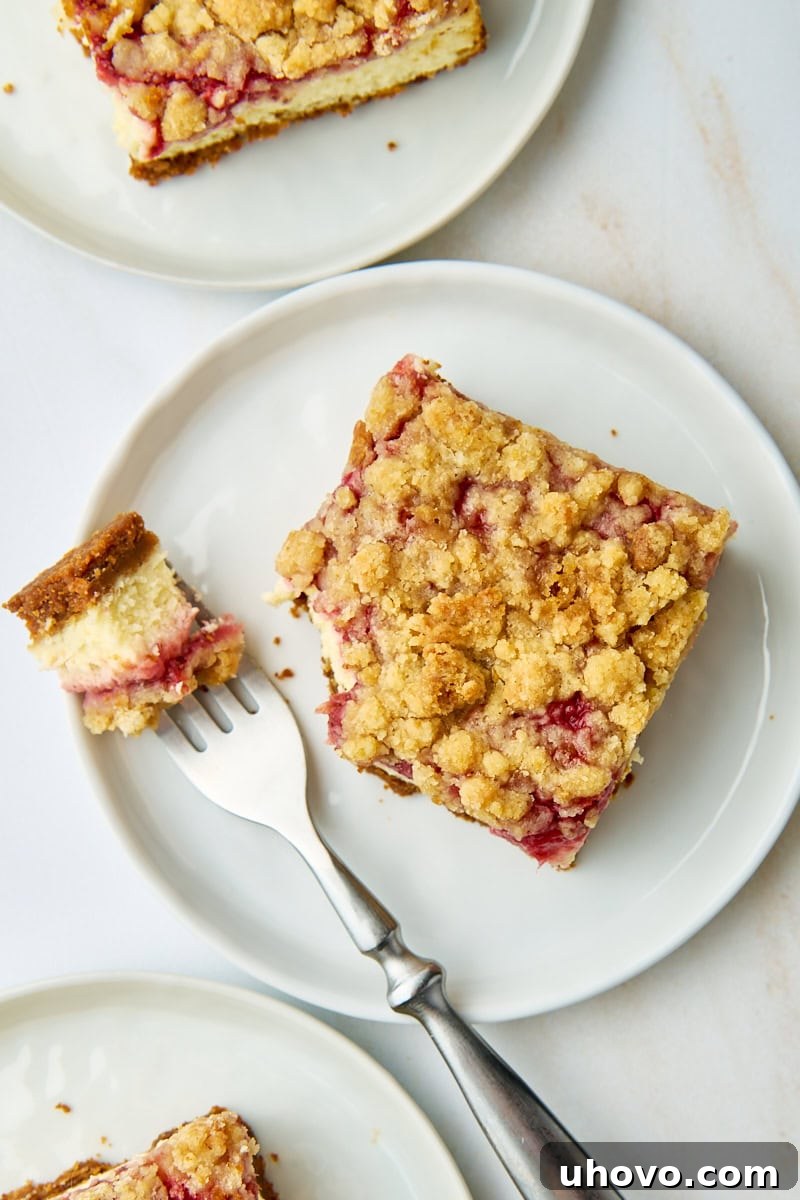 Overhead close-up of a delightful strawberry cheesecake bar on a white plate with a perfect bite taken out by a fork, with more servings partially visible in the blurred background.