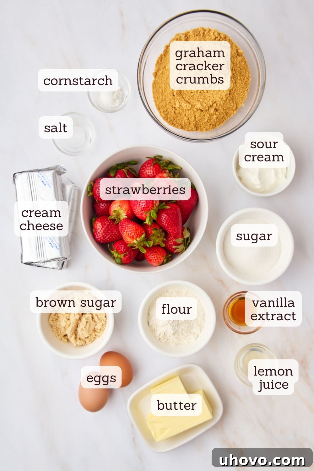 Overhead view of fresh ingredients laid out for making delicious strawberry cheesecake bars, including graham crackers, blocks of cream cheese, a carton of sour cream, and a bowl of vibrant red strawberries.