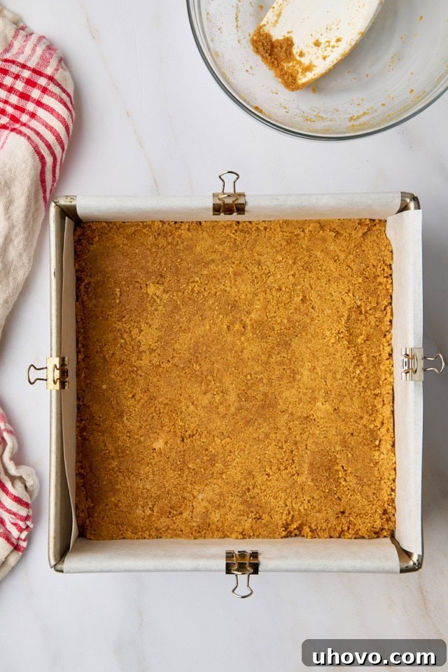 Top-down view of the graham cracker crust perfectly pressed into the bottom of a square baking pan, forming an even layer before baking.