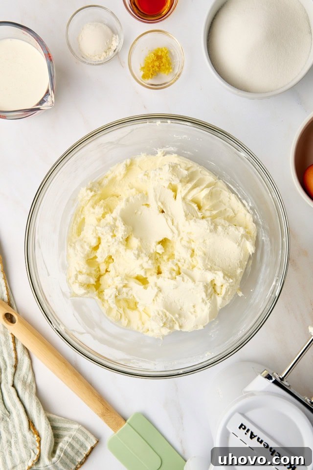 An overhead shot shows softened cream cheese being beaten to a smooth consistency in a clear glass mixing bowl, a crucial first step for a lump-free cheesecake.