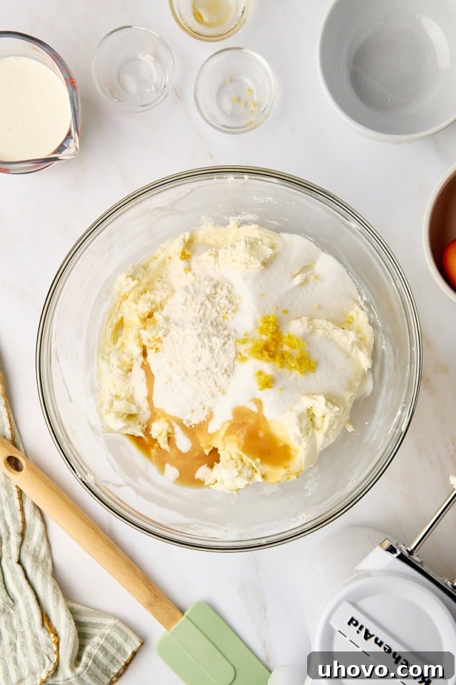 An overhead view displays granulated sugar, vanilla extract, all-purpose flour, and fresh lemon zest being carefully added to the already beaten cream cheese mixture in a large glass bowl.