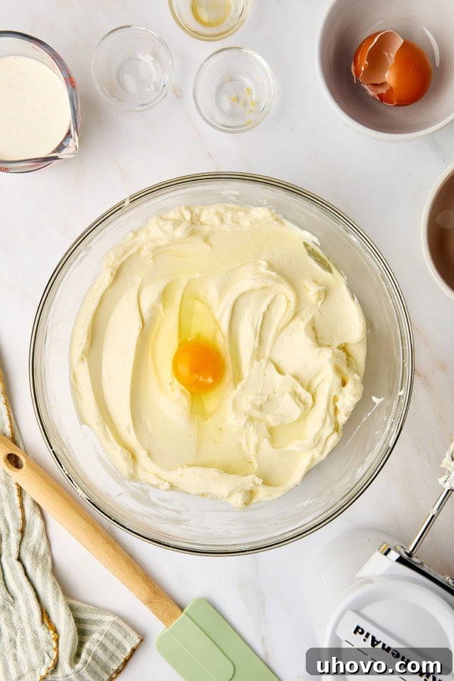 An overhead perspective as a single egg is gently cracked and added to the cheesecake mixture, ensuring it's incorporated one at a time.