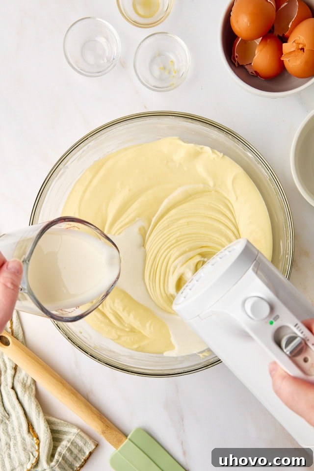 An overhead view shows a steady stream of heavy cream being poured into the cheesecake batter, ready to be gently mixed in for a perfect consistency.