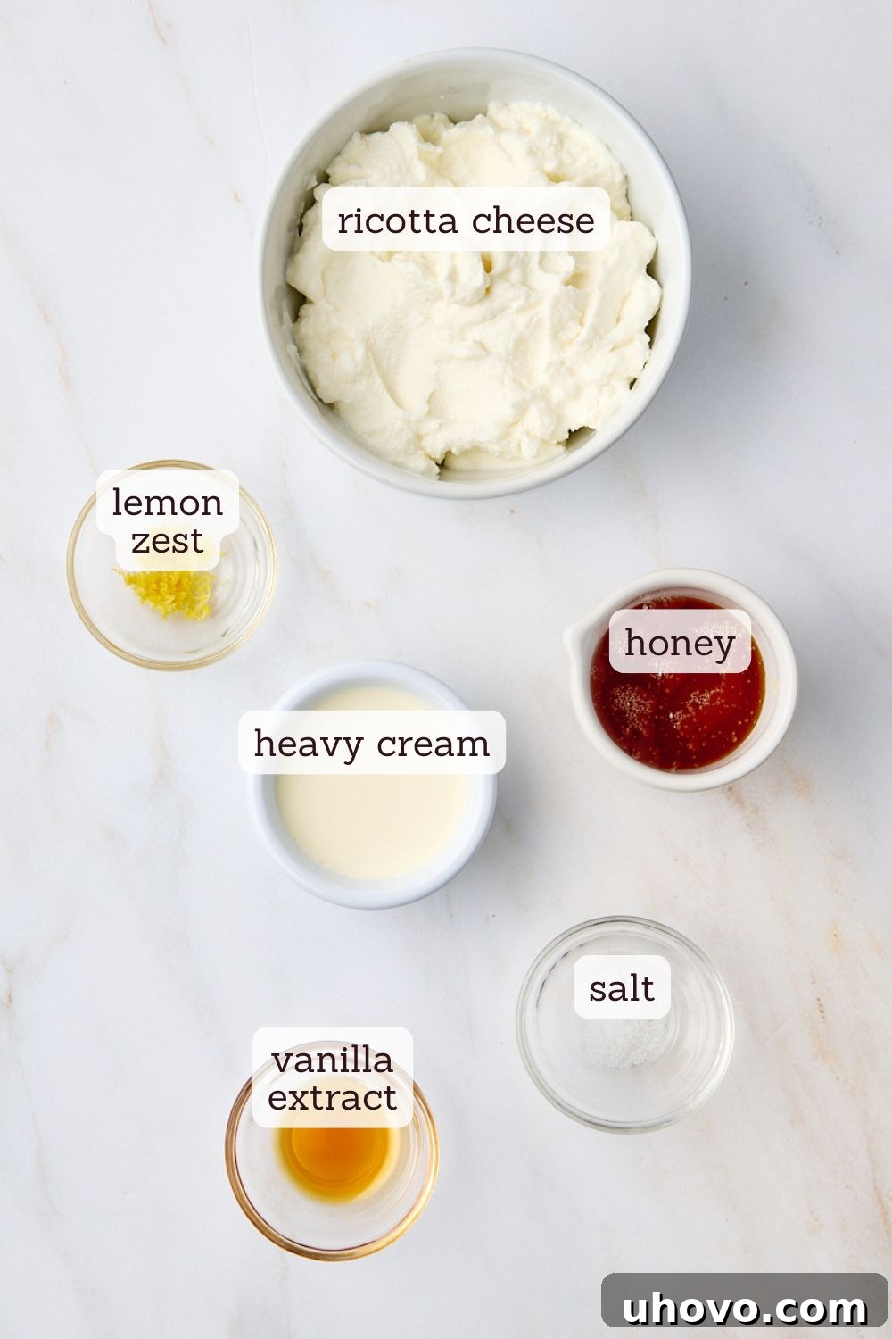 Overhead view of the essential ingredients for making ricotta mousse laid out on a clean surface: a bowl of smooth ricotta cheese, a jar of golden honey, a bottle of vanilla extract, a zester with fresh lemon zest, and a small pinch of salt.