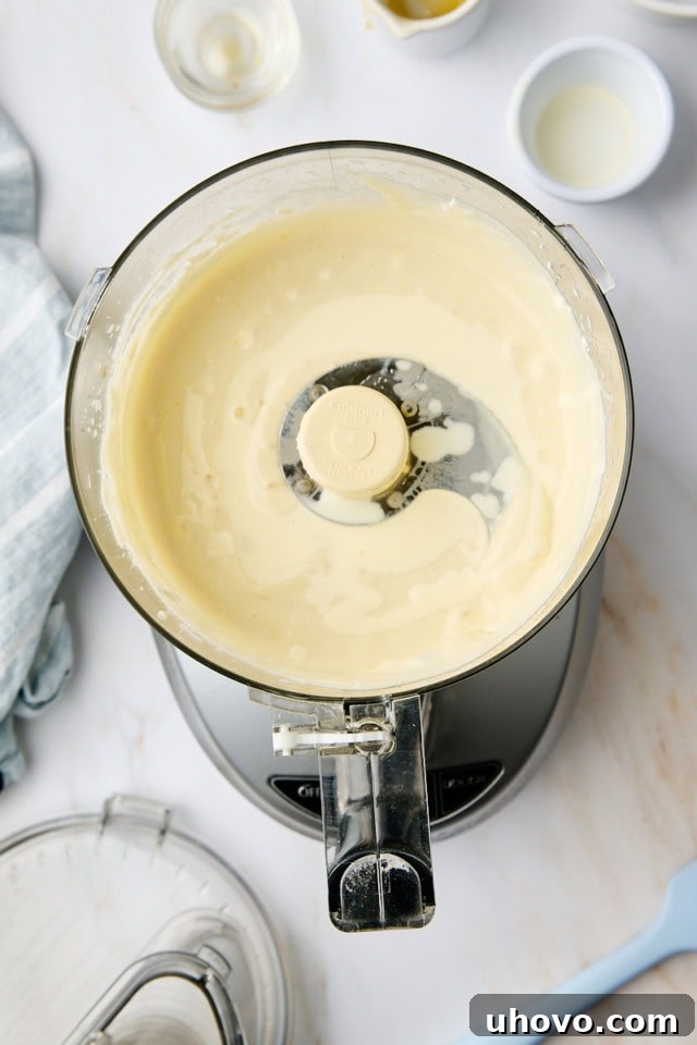 A stream of heavy cream is being poured into the food processor bowl, which already contains the blended ricotta mousse mixture, preparing for the final blending step.