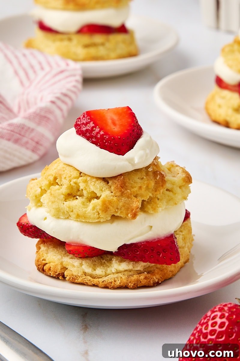 A beautifully assembled strawberry shortcake on a pristine white plate, with two more shortcakes softly blurred in the background, highlighting its delightful layers of biscuit, strawberries, and whipped cream.