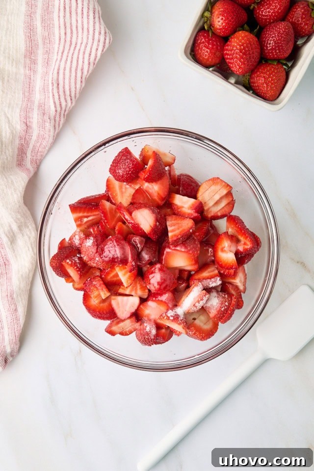 An overhead view of a glass mixing bowl filled with sliced strawberries, granulated sugar, and fresh lemon juice, illustrating the process of macerating the berries for the shortcake filling.