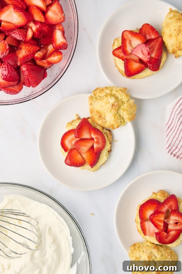 A dynamic overhead shot showing the assembly process of strawberry shortcakes, with biscuits split, topped with macerated strawberries, and a dollop of whipped cream, ready for the top biscuit layer.