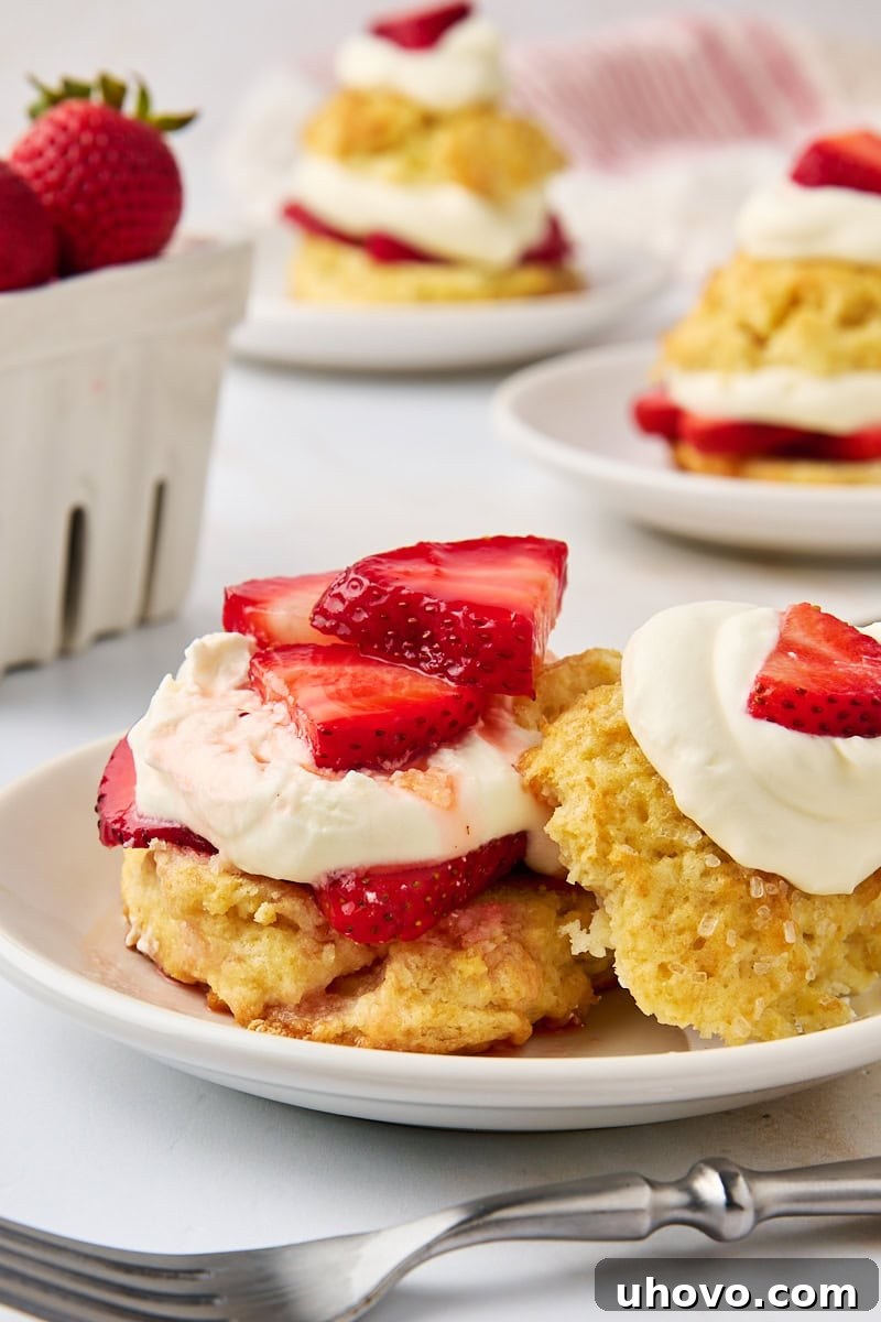 A close-up view of a strawberry shortcake on a white plate, with the top biscuit gently set aside, revealing the luscious layers of strawberries and whipped cream beneath. More shortcakes and a basket of strawberries are softly blurred in the background.