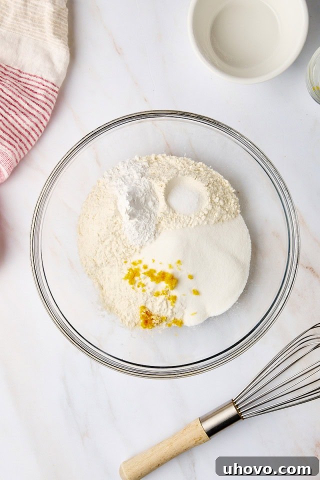 An overhead view of a glass mixing bowl containing flour, sugar, baking powder, salt, and lemon zest, illustrating the first step of combining dry ingredients for the shortcake biscuits.