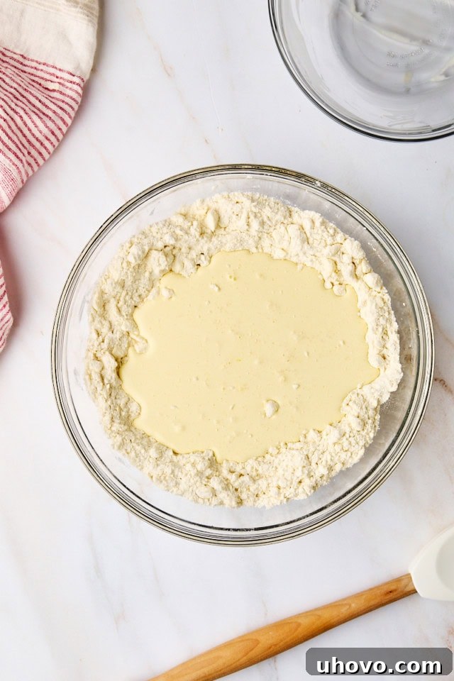 An overhead shot depicting wet ingredients (cream, egg, vanilla) being poured into the dry flour and butter mixture, indicating the next phase of biscuit dough preparation.