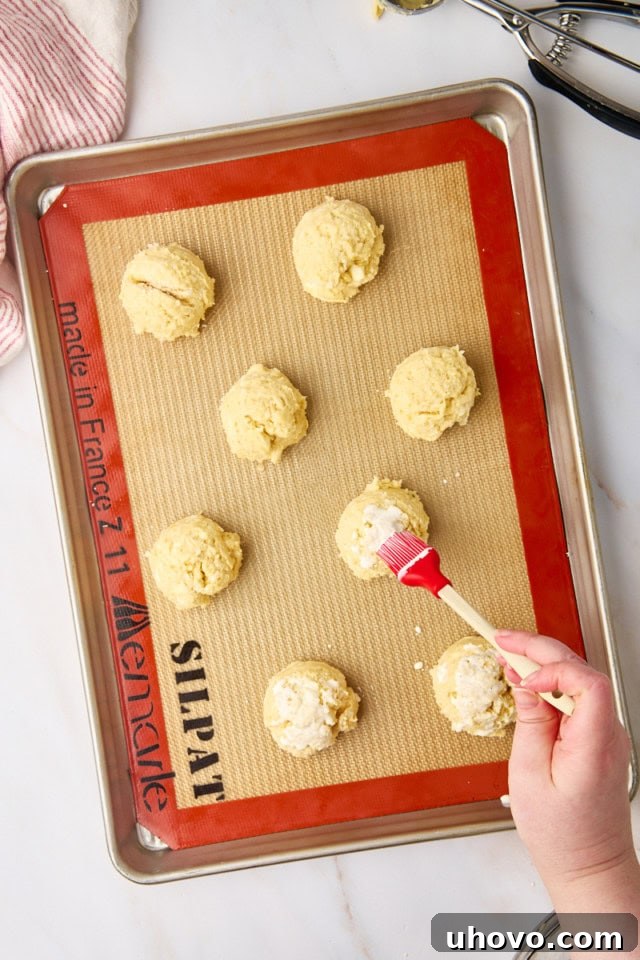 An overhead view of dollops of biscuit dough arranged on a lined baking sheet, with a silicone brush applying cream to their tops, and coarse sugar sprinkled over, ready for baking.