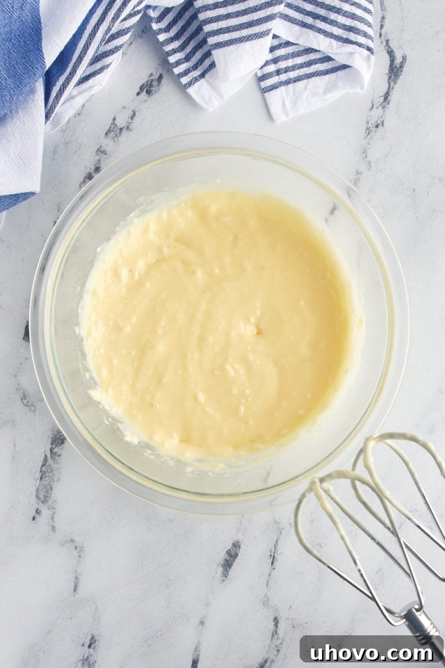 Overhead view of the smooth and creamy mixture of softened cream cheese, sugar, egg, and lemon juice in a glass mixing bowl, prepared for the topping.