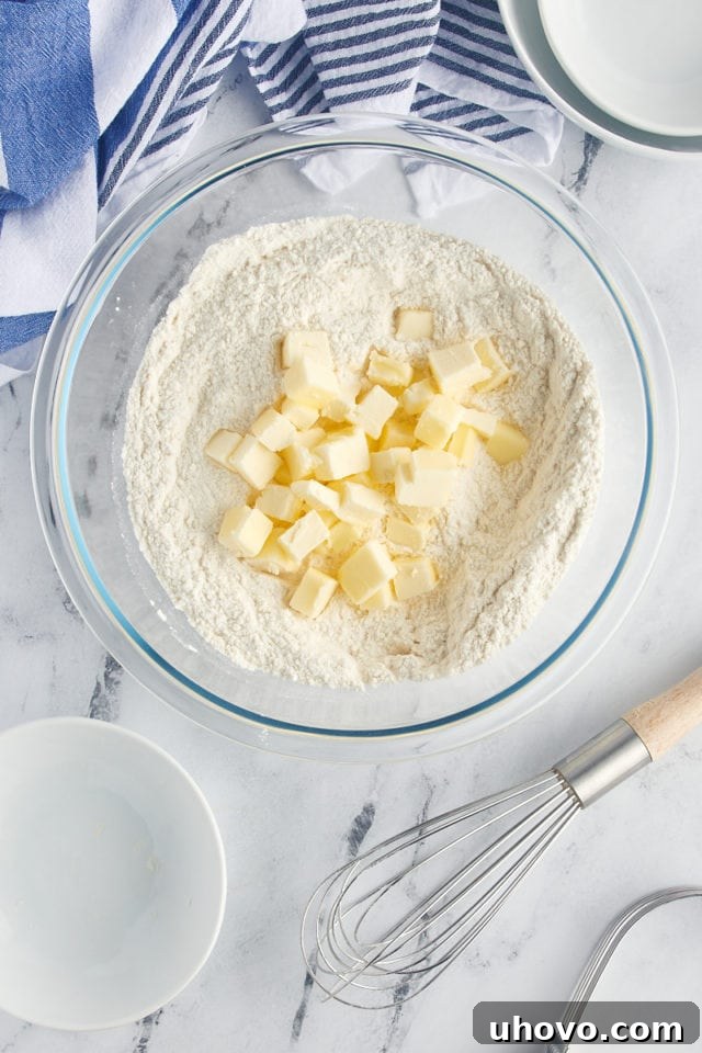 Cold, cubed unsalted butter being added to the flour and sugar mixture in a mixing bowl, prior to being cut in for a crumbly texture.